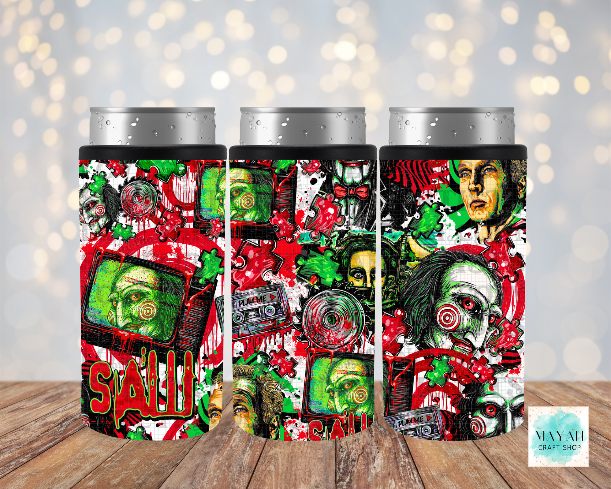 Three insulated can coolers with silver aluminum can tops, featuring a colorful horror-themed collage print of a face, cassette tapes, displayed on a wooden surface against a blurred bokeh background.