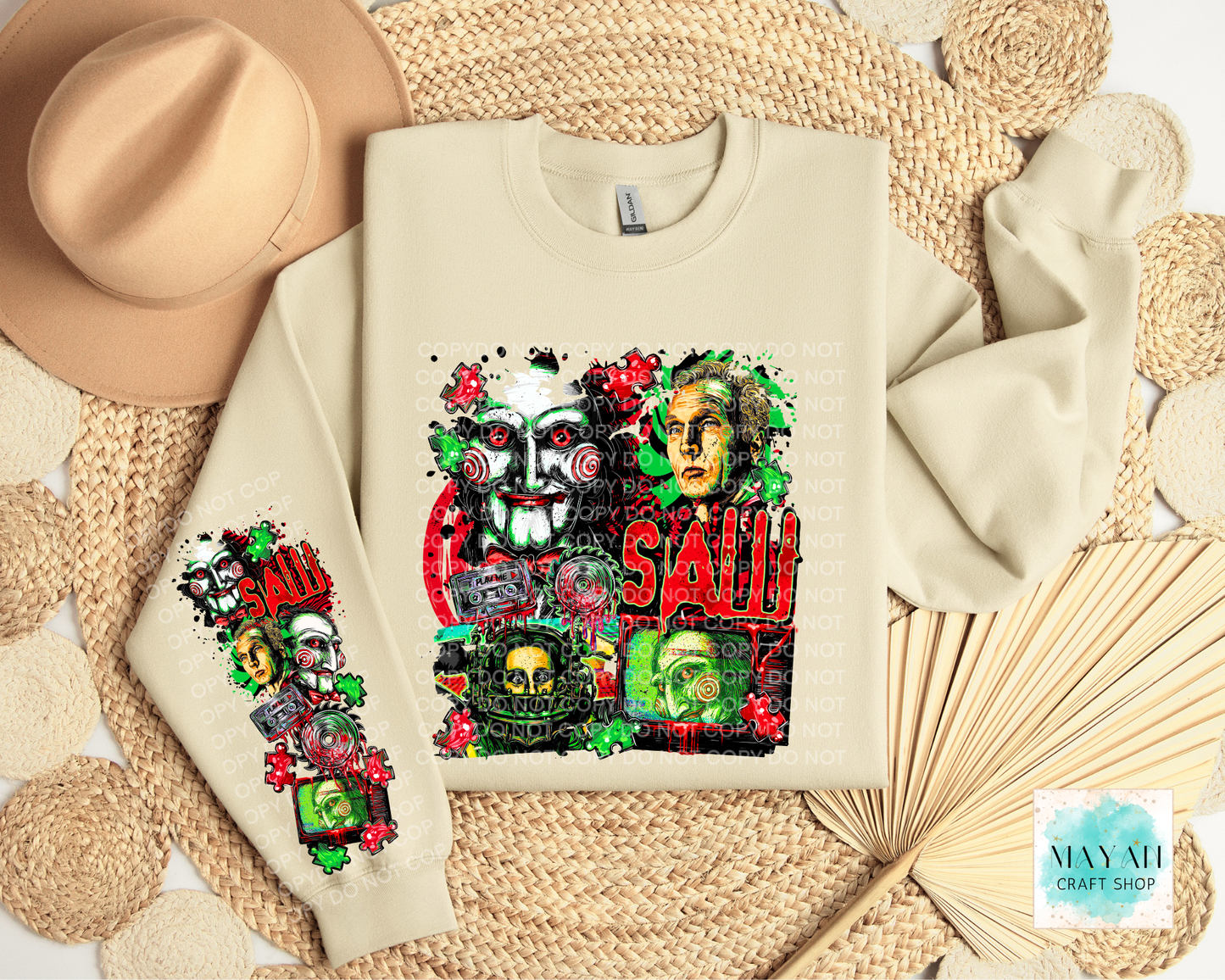 A cream-colored crewneck sweatshirt featuring a detailed graphic print of the character on the front and sleeve, styled in a flat-lay arrangement with a beige hat, woven placemats, and a dried palm leaf fan.
