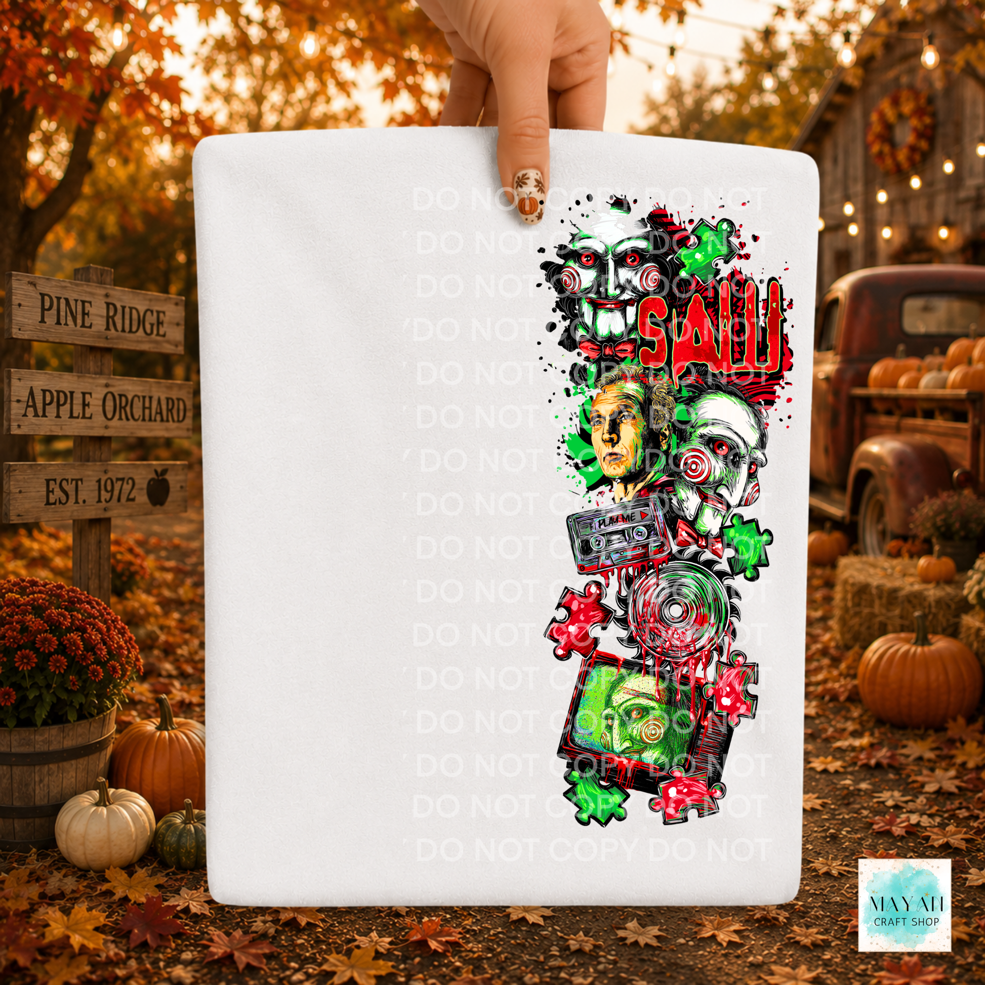 A hand holds a white rectangular fabric item, likely a bandana or small flag, featuring a colorful graphic print of horror-themed skulls, a cassette tape, puzzle pieces. The background is a blurred autumn outdoor scene with pumpkins, fall leaves, and a rustic truck.