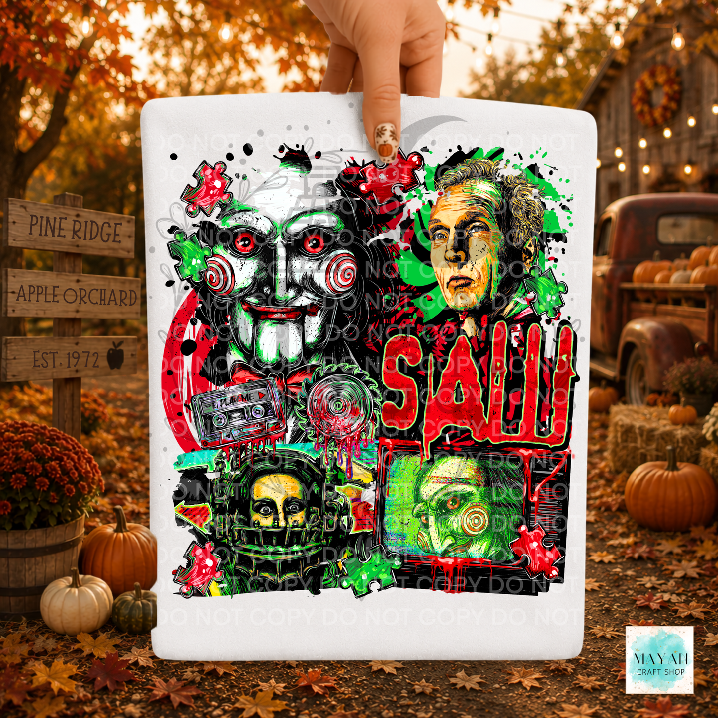 A hand holds a white shirt featuring a horror-themed collage with multiple illustrated faces, red lettering, and green/red paint splatter details. The background is a fall outdoor scene with pumpkins, autumn leaves, and rustic signs.