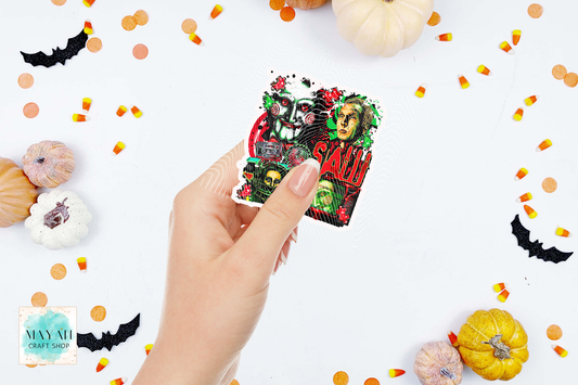 A hand holds a square Halloween-themed sticker featuring a skull, a green monster face, and a word in red. The background is white and decorated with small pumpkins, candy corn, orange confetti, and black bat cutouts.