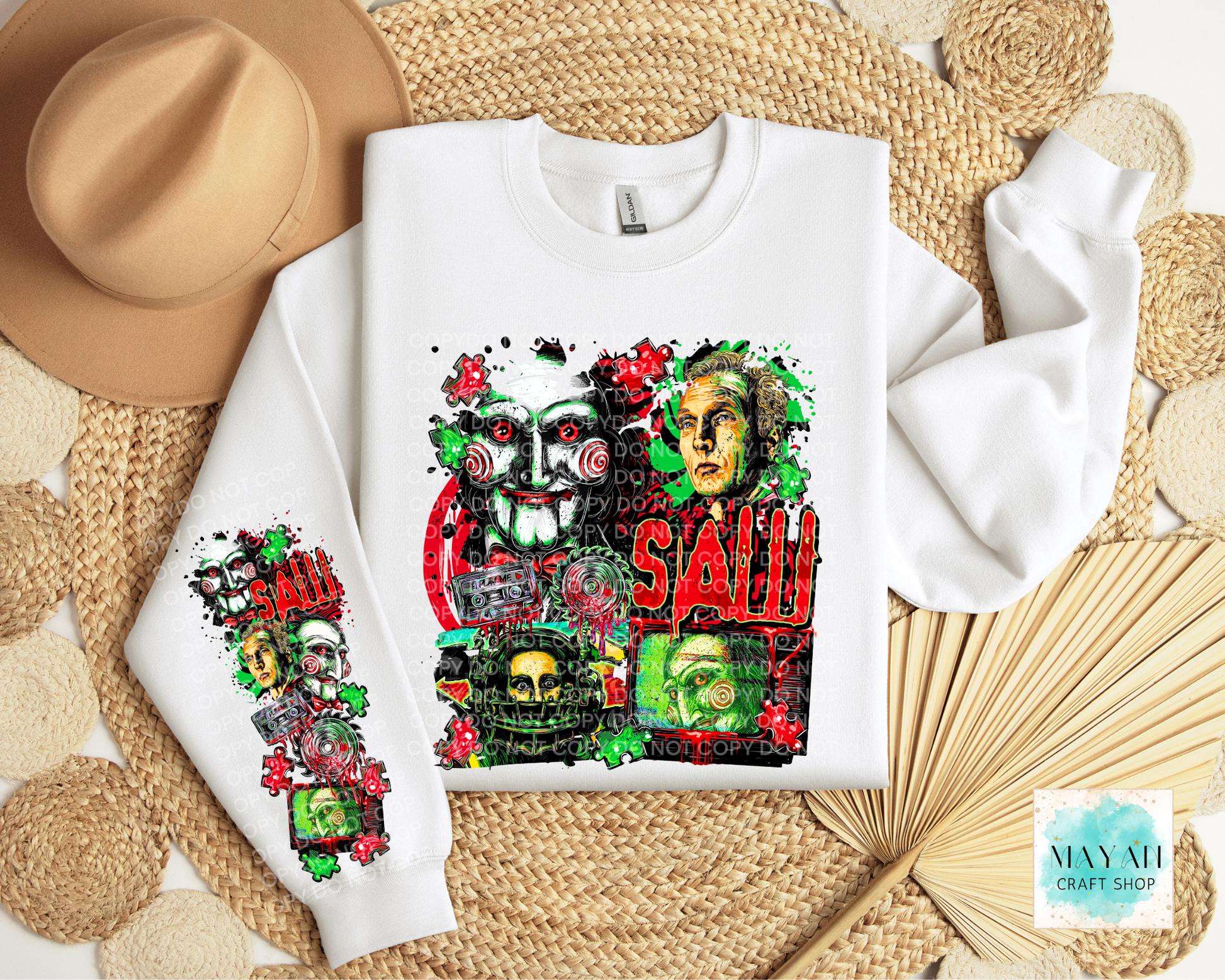 A white crewneck sweatshirt with a large horror-themed graphic print on the front and sleeve, styled in a flat lay with a tan hat, woven mat, and paper fan.
