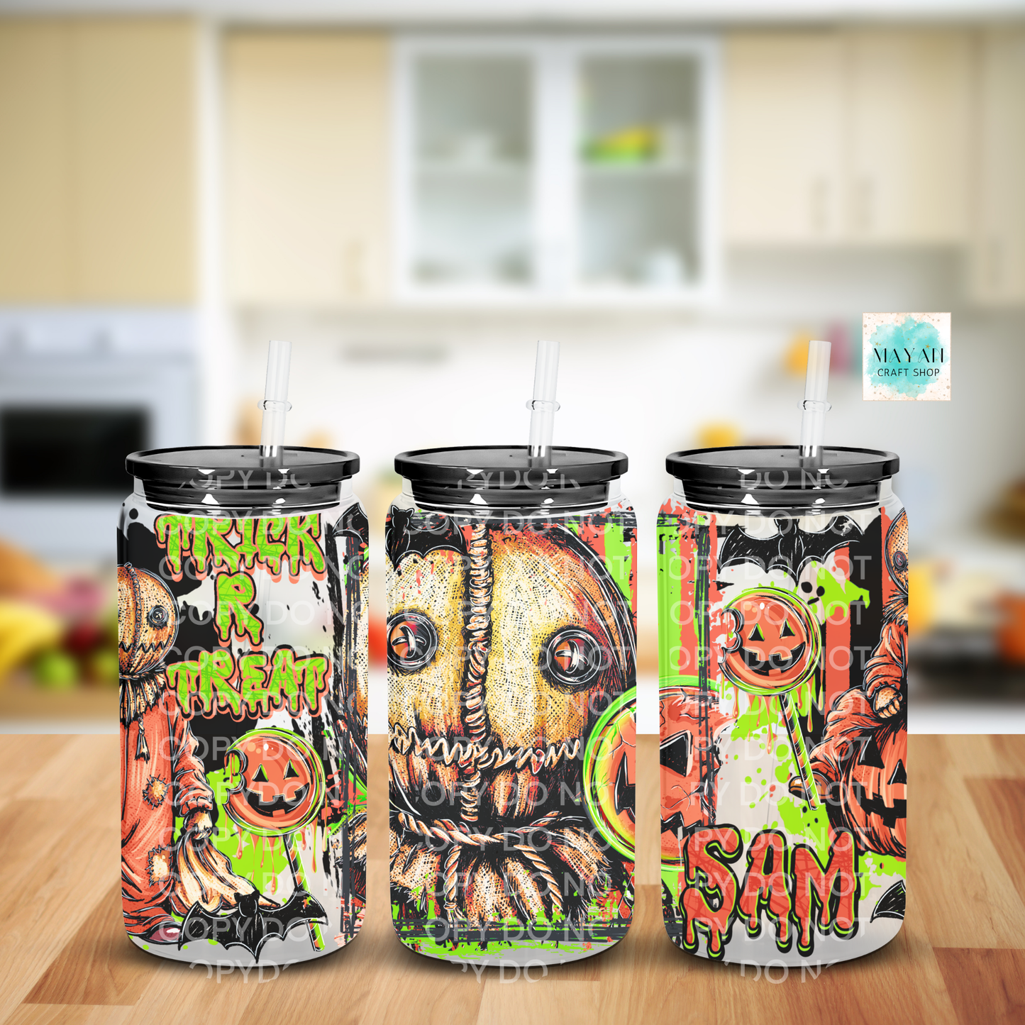 Three tall frosted glass cans with black lids and clear straws, decorated with Halloween-themed graphics including pumpkins, ghosts, and the words 'TRICK or TREAT', shown on a wooden surface with a blurred kitchen background.