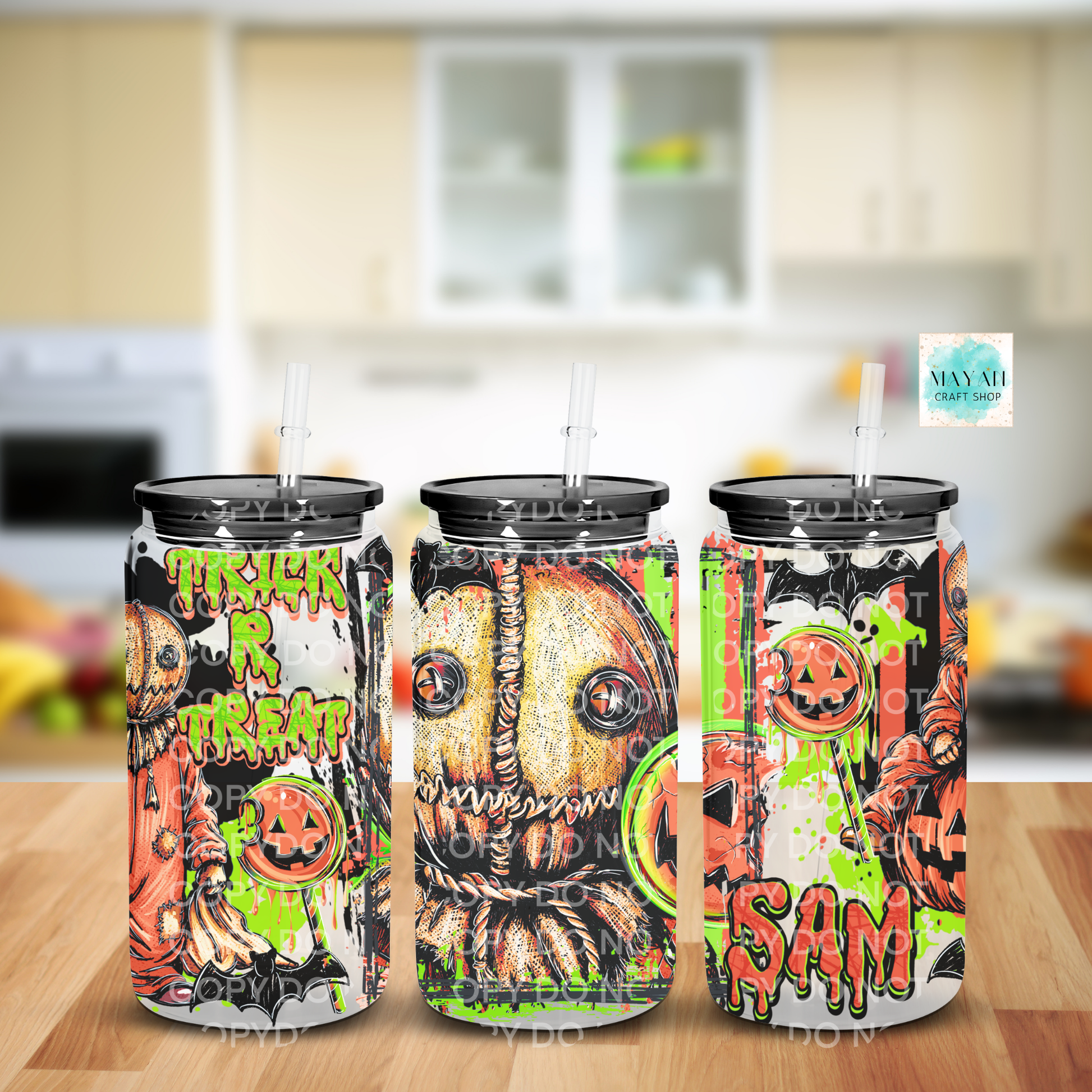 Three tall frosted glass cans with black lids and clear straws, decorated with Halloween-themed graphics including pumpkins, ghosts, and the words 'TRICK or TREAT', shown on a wooden surface with a blurred kitchen background.