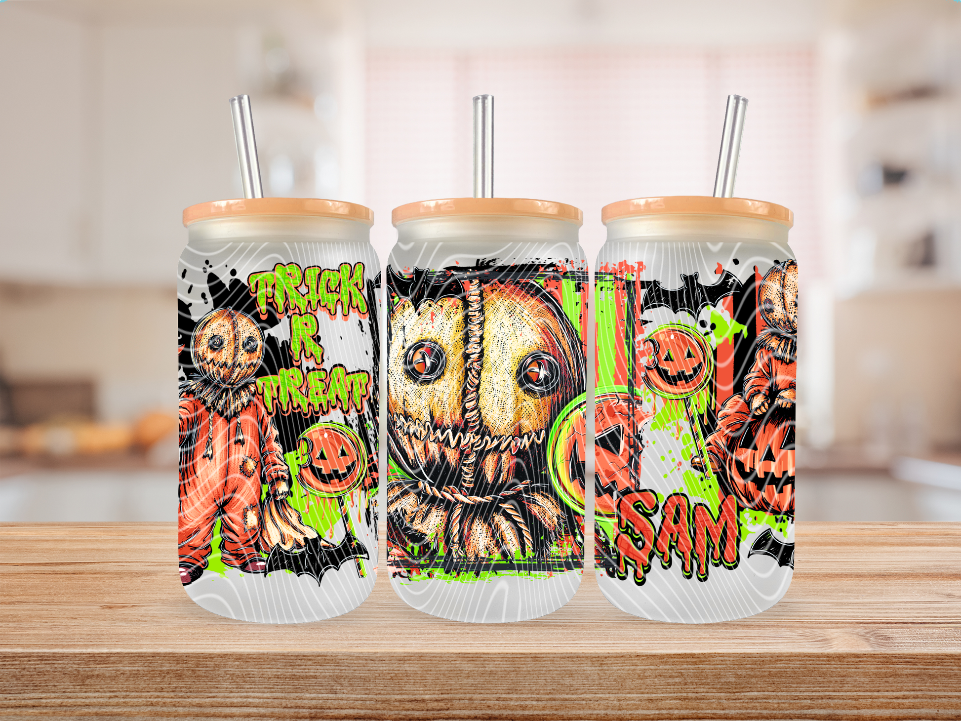 Three frosted glass cans with light wood lids and metal straws, decorated with a spooky Halloween graphic featuring pumpkins, a stitched face, displayed on a wooden surface with a blurred kitchen background.