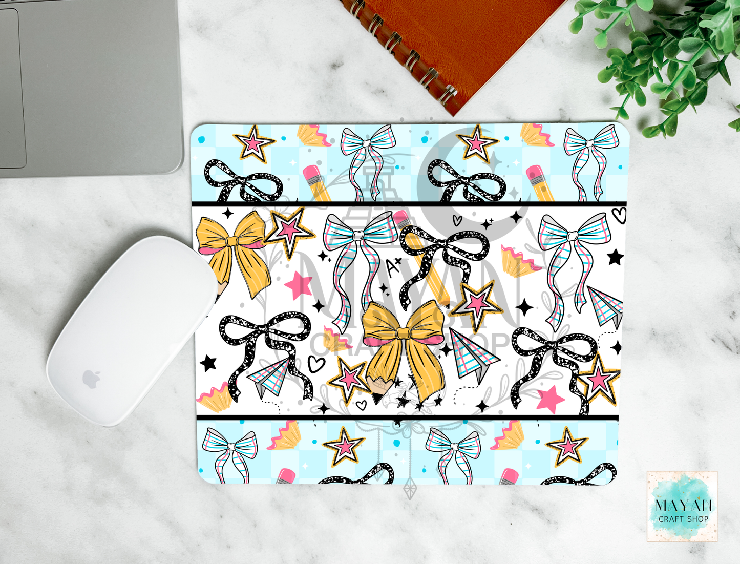 A mouse pad with a colorful pattern of bows, stars, pencils, and kites, styled on a white marble surface next to a white computer mouse, a silver laptop corner, an orange spiral notebook, and a green plant.