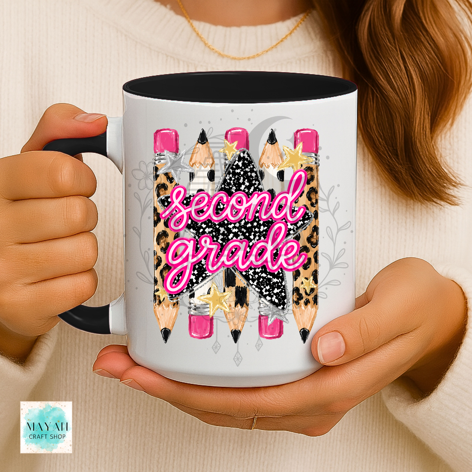 A person holds a white ceramic mug with a black handle and interior. The mug features a 'second grade' birthday graphic with pink script text, leopard print pencils, and glittery stars.