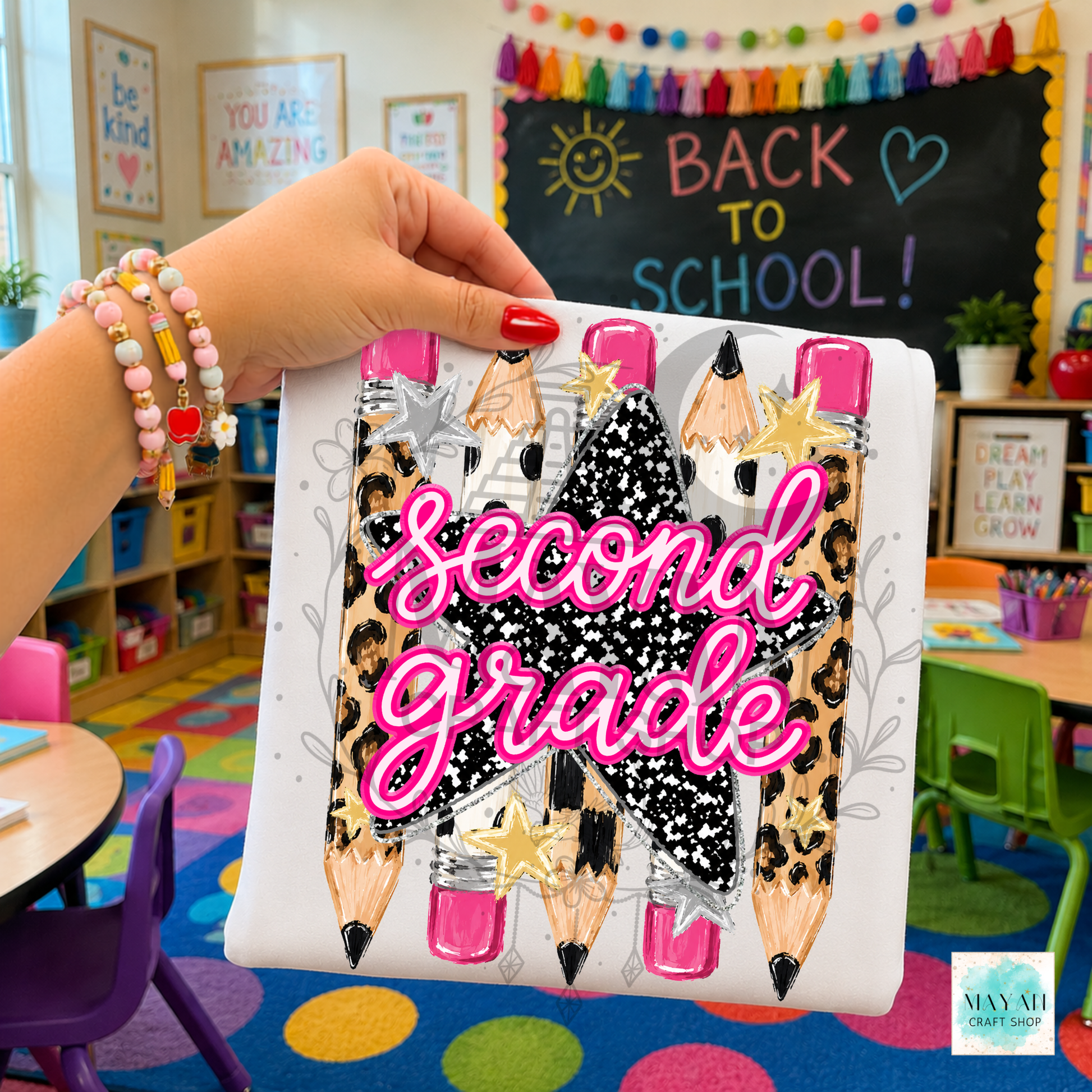A hand holds a white shirt featuring a decorative school-themed design with the words 'second grade' in pink script over illustrated pencils and a black star. The background shows a colorful children's classroom with a chalkboard reading 'BACK TO SCHOOL!'.