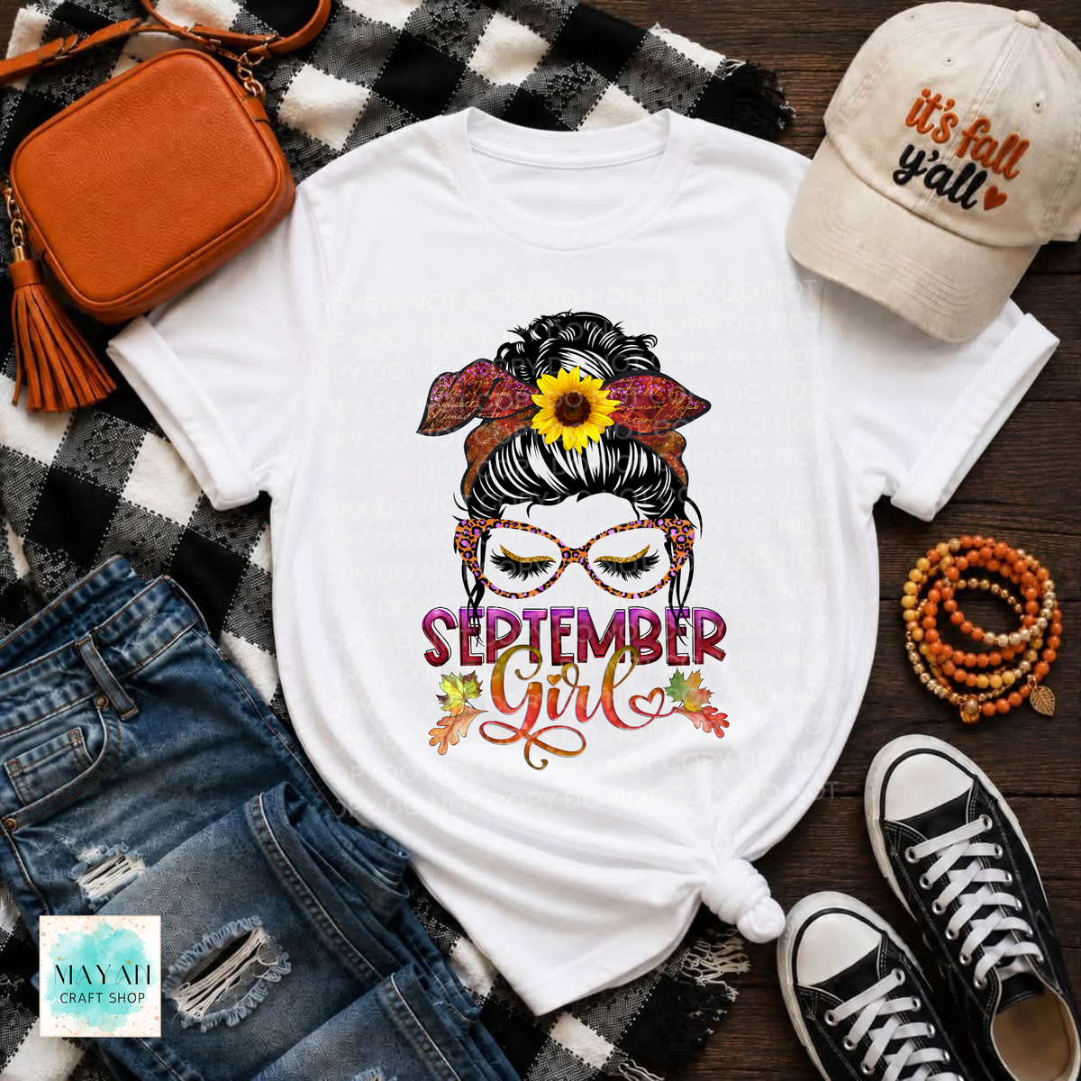 A white graphic T-shirt laid flat on a black-and-white plaid blanket with jeans, an orange crossbody bag, a beige cap reading 'it's fall y'all', beaded bracelets, and black-and-white sneakers around it. The shirt features a stylized woman with glasses, a sunflower, leaves, and the words 'SEPTEMBER Girl'.