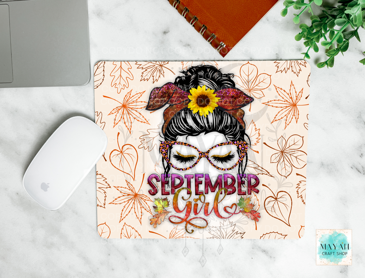 A rectangular desk mat featuring a 'September Girl' illustration with glasses, a sunflower in her hair, and autumn leaf patterns, shown in a styled flat-lay on a white marble surface with a laptop, mouse, notebook, and greenery.