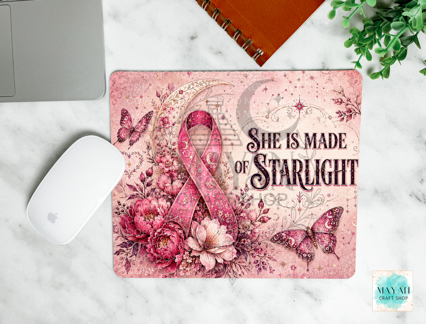 A rectangular mouse pad with a pink floral and ribbon design sits on a white marble surface. The mat features the text 'She Is Made Of Starlight' with butterflies, flowers, and a crescent moon.