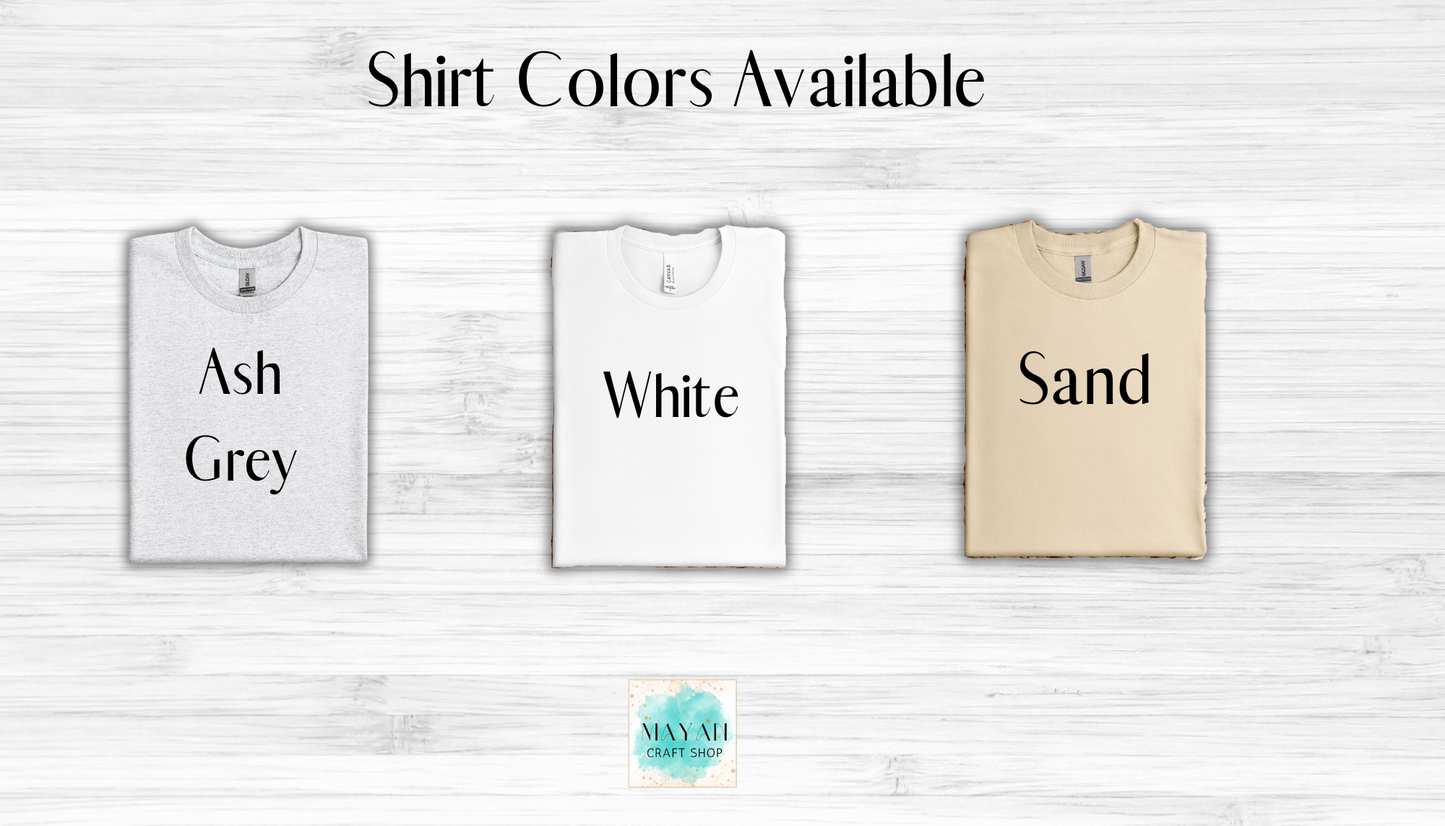 Three t-shirts in ash grey, white, and sand with text labels on a light wooden background.