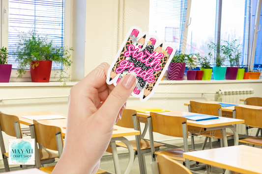 A hand holds a die-cut sticker featuring pink glitter-style text over a leopard print and pencil illustration, with a blurred classroom background.