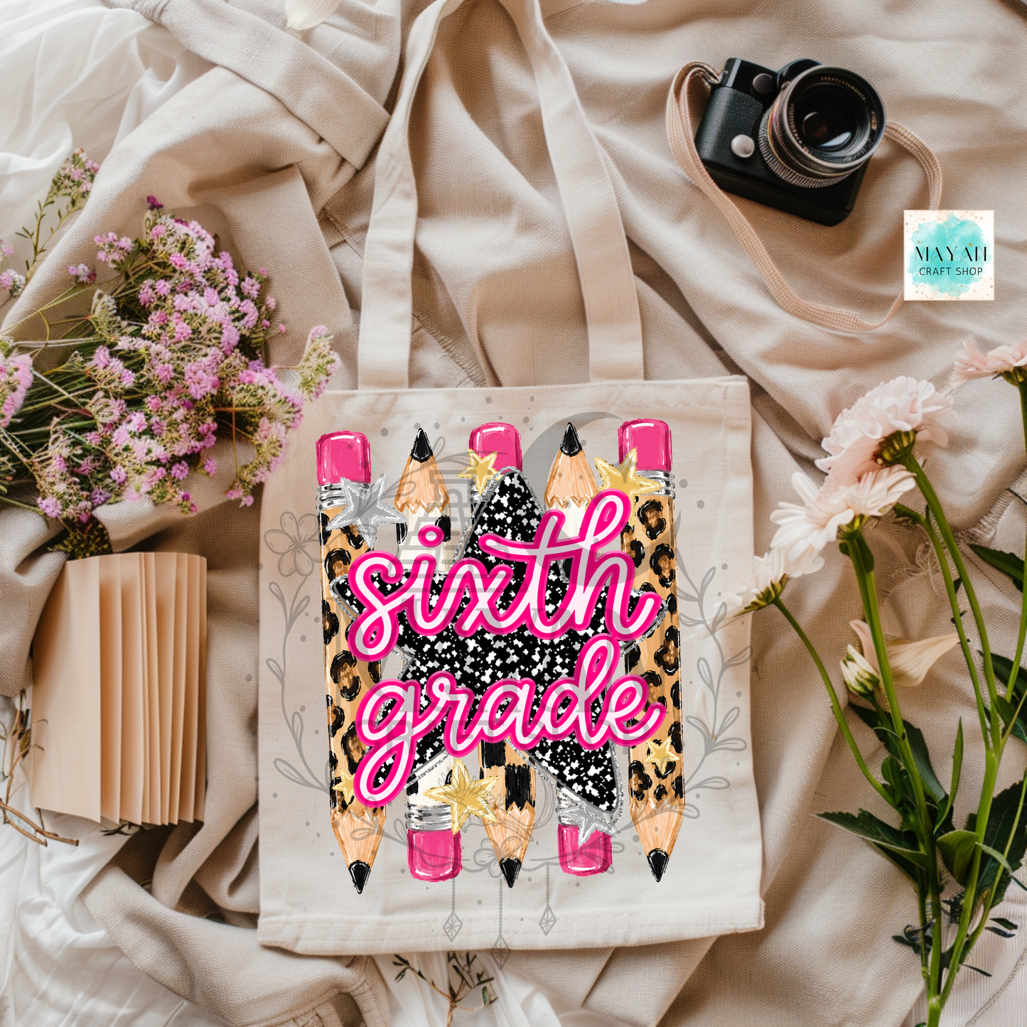 A canvas tote bag with a 'sixth grade' school-themed graphic featuring cartoon pencils and leopard print, styled in a flat lay with flowers, a camera, and a notebook on beige fabric.