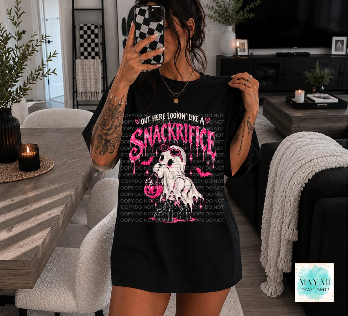 A woman takes a mirror selfie in a modern living room while wearing an oversized black graphic T-shirt. The shirt features a pink and white ghost illustration with the text 'OUT HERE LOOKIN' LIKE A SNACKRITFICE' and a repeating 'DO NOT COPY' watermark pattern.