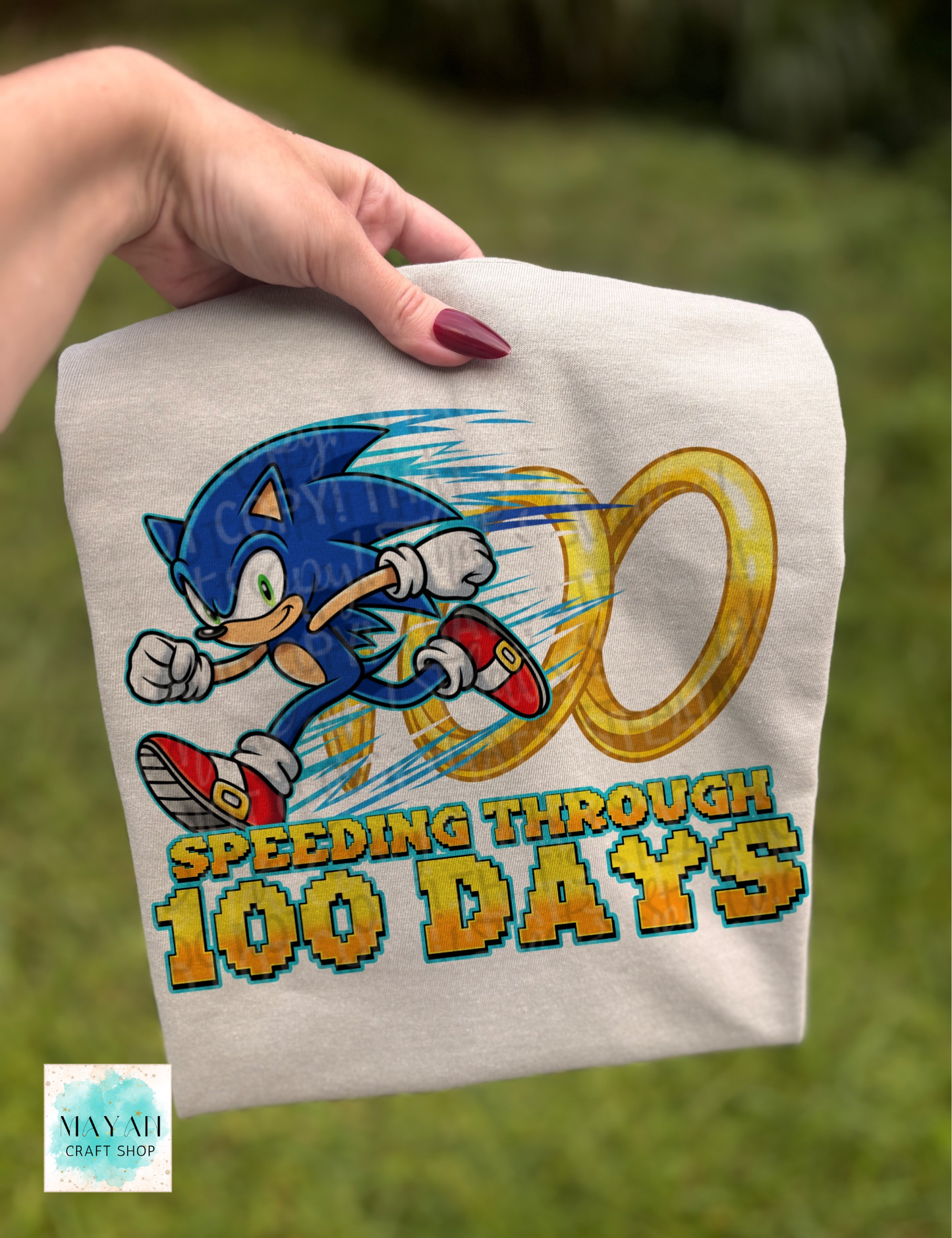 T-shirt with Sonic the Hedgehog design and text 'Speeding Through 100 Days' held by a hand against a grassy background.