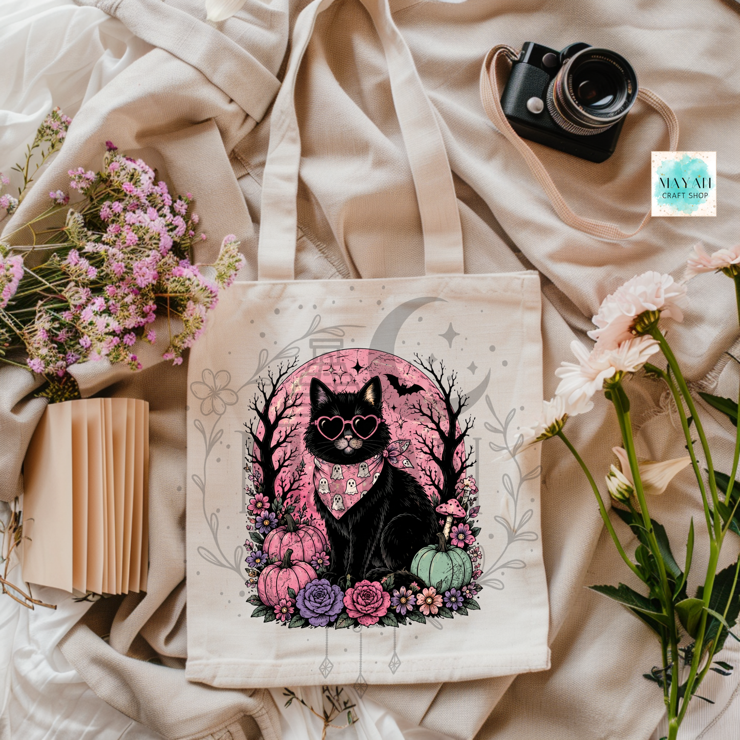 Tote bag with a black cat design on a beige fabric background with flowers and a camera.