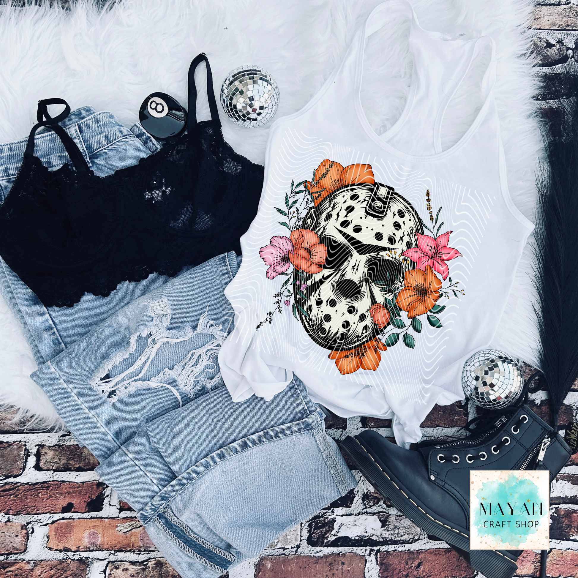 White tank top with skull and floral design on a brick surface, surrounded by denim shorts and black lace top.
