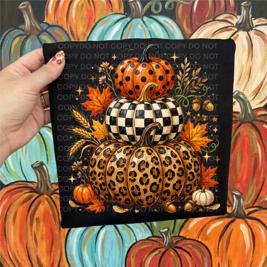 A hand holds a black shirt featuring a stacked autumn pumpkin illustration with orange, black-and-white checkered, and leopard-print pumpkins, maple leaves, wheat, and acorns. The background is a painted scene of colorful pumpkins.