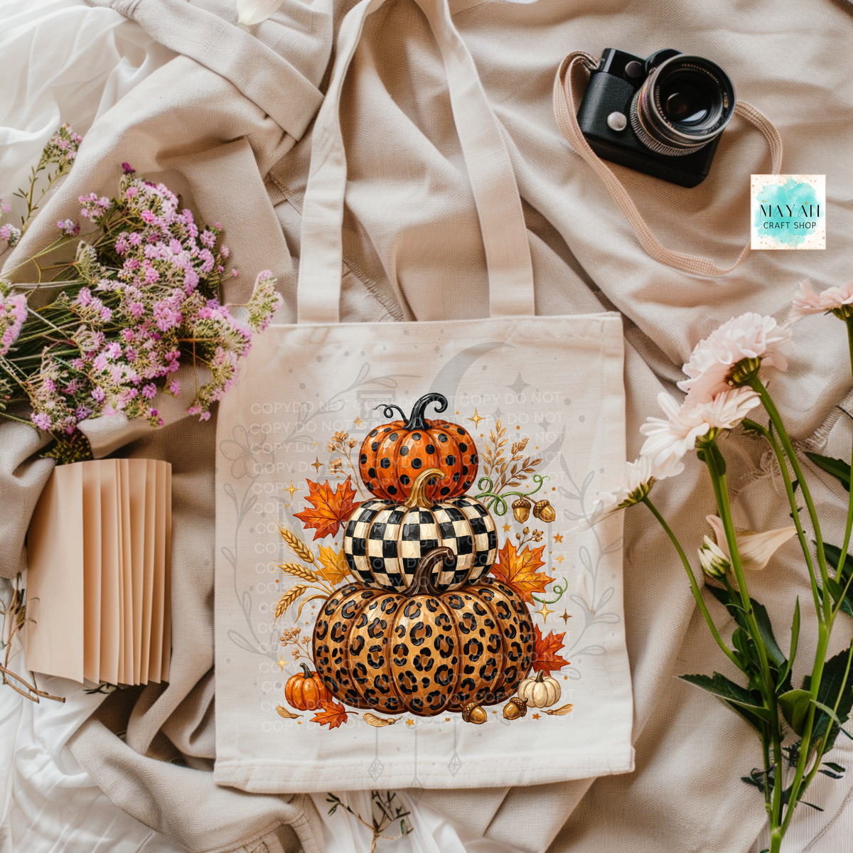 A cream canvas tote bag with a stacked autumn pumpkin graphic featuring leopard print, checkered pattern, and orange pumpkins, styled in a flat lay with flowers, a camera, and beige fabric.