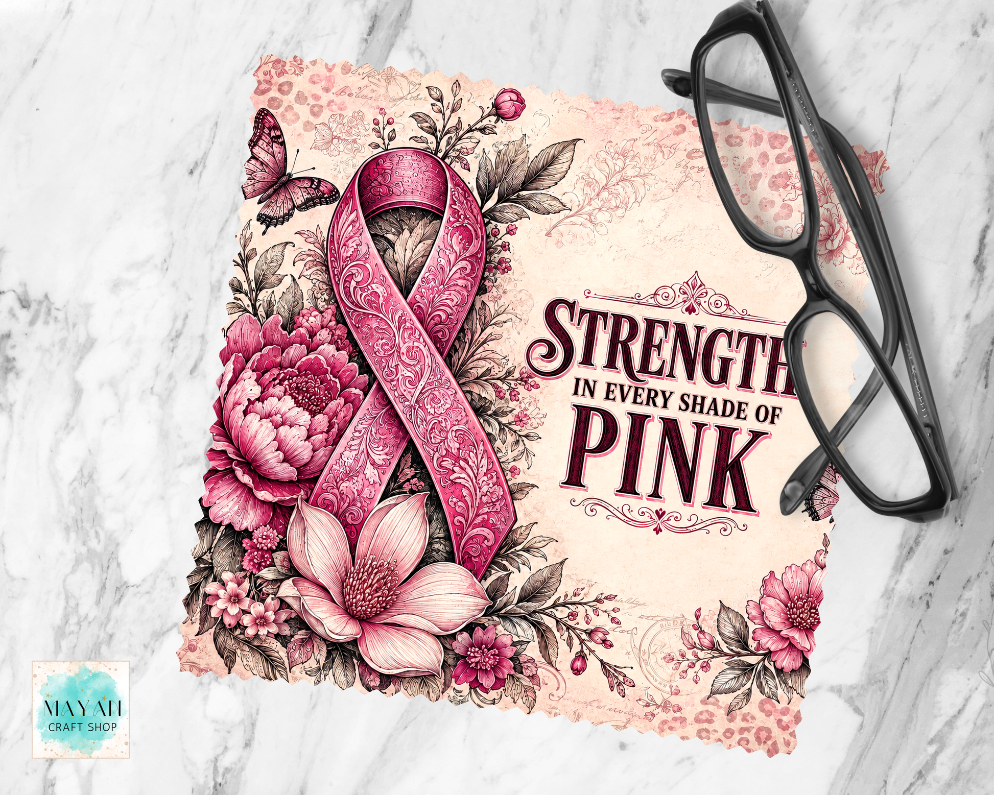 A lens cloth cleaner featuring a pink patterned awareness ribbon, pink flowers, leaves, and the text 'STRENGTH IN EVERY SHADE OF PINK,' styled on a light marble surface with black eyeglasses placed on top. 