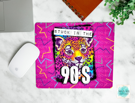 A rectangular mouse pad with a vibrant 90s aesthetic, featuring a colorful leopard graphic and the text 'STUCK IN THE 90S' on a pink background with neon patterns. The mouse pad is styled in a flat-lay on a white marble desk alongside a white computer mouse, a laptop, a notebook, and a small plant.