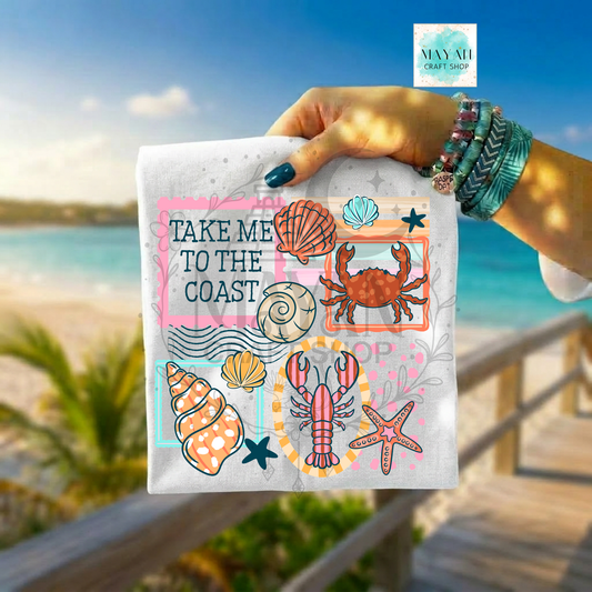 Hand holding a shirt with coastal designs and text 'Take Me to the Coast' against a beach background.