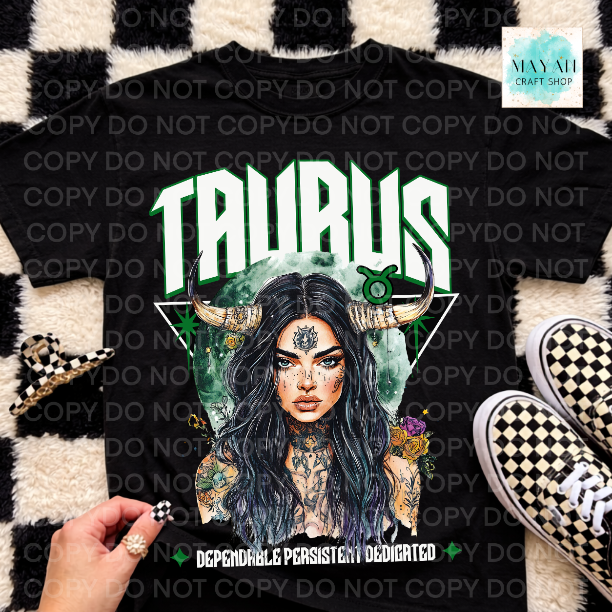 A black graphic t-shirt featuring a detailed Taurus zodiac design with a horned woman, green text, and a green astrological symbol. The shirt is styled in a flat-lay arrangement on a black and white checkered rug with a hand holding a matching checkered accessory, alongside sneakers.