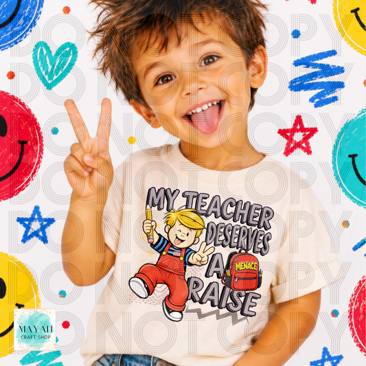 A young boy with brown hair smiling and sticking out his tongue, making a peace sign. He is wearing a cream-colored t-shirt with a cartoon graphic of a boy in red overalls and the text 'MY TEACHER DESERVES A RAISE'.