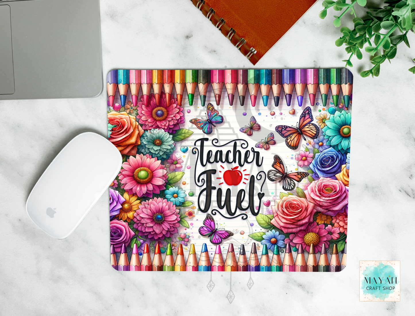 A mouse pad with a colorful 'Teacher Fuel' design featuring flowers, butterflies, and a row of colored pencils, shown in a flat-lay on a white marble surface next to a white computer mouse and an orange notebook.