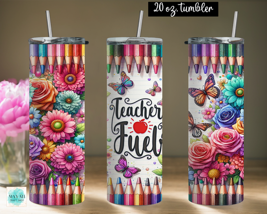Three tall skinny tumblers with clear straws are shown on a tabletop against a warm beige wall. The tumblers feature colorful printed artwork: rows of colored pencils at the top and bottom, large flowers, butterflies, and one center design with the words 'Teacher Fuel' and a red apple.