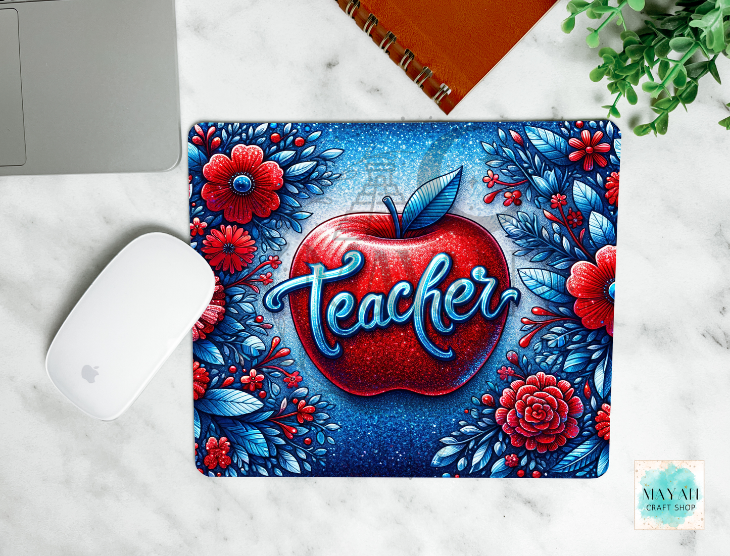 A mouse pad with a vibrant 'Teacher' apple design, shown in a flat-lay arrangement on a white marble surface alongside a white computer mouse, a brown notebook, and a small green plant.