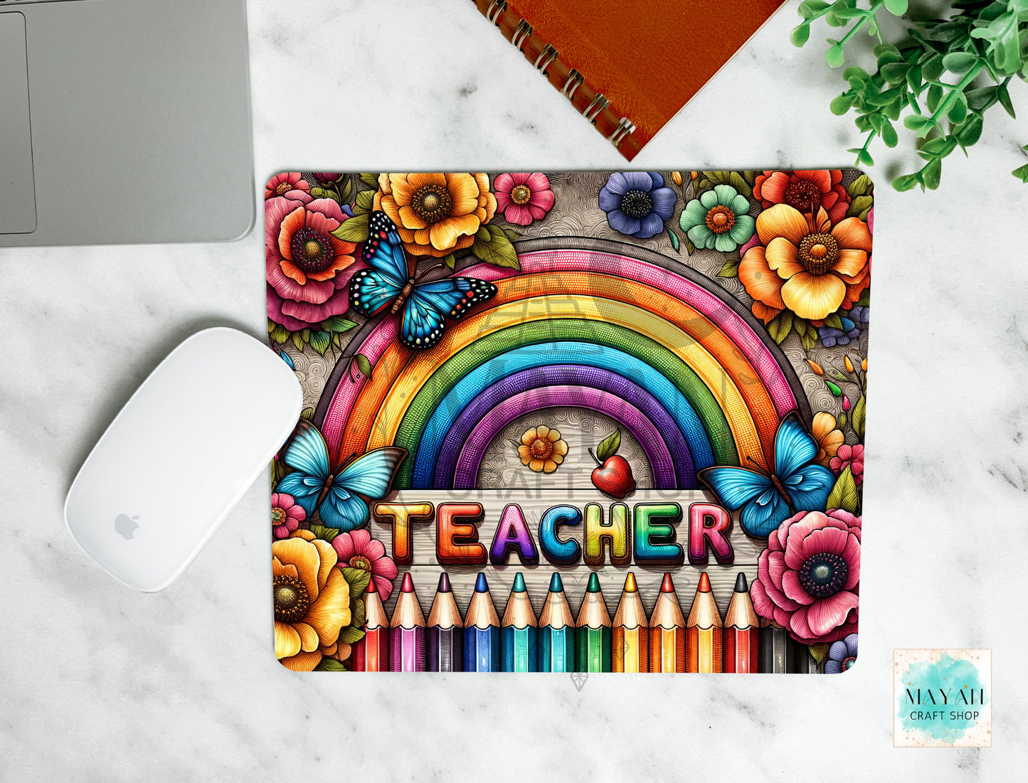 A mouse pad featuring a rainbow, flowers, butterflies, colored pencils, and the word 'TEACHER' in raised letters, shown on a white marble surface with a laptop mouse, notebook, and plant nearby.