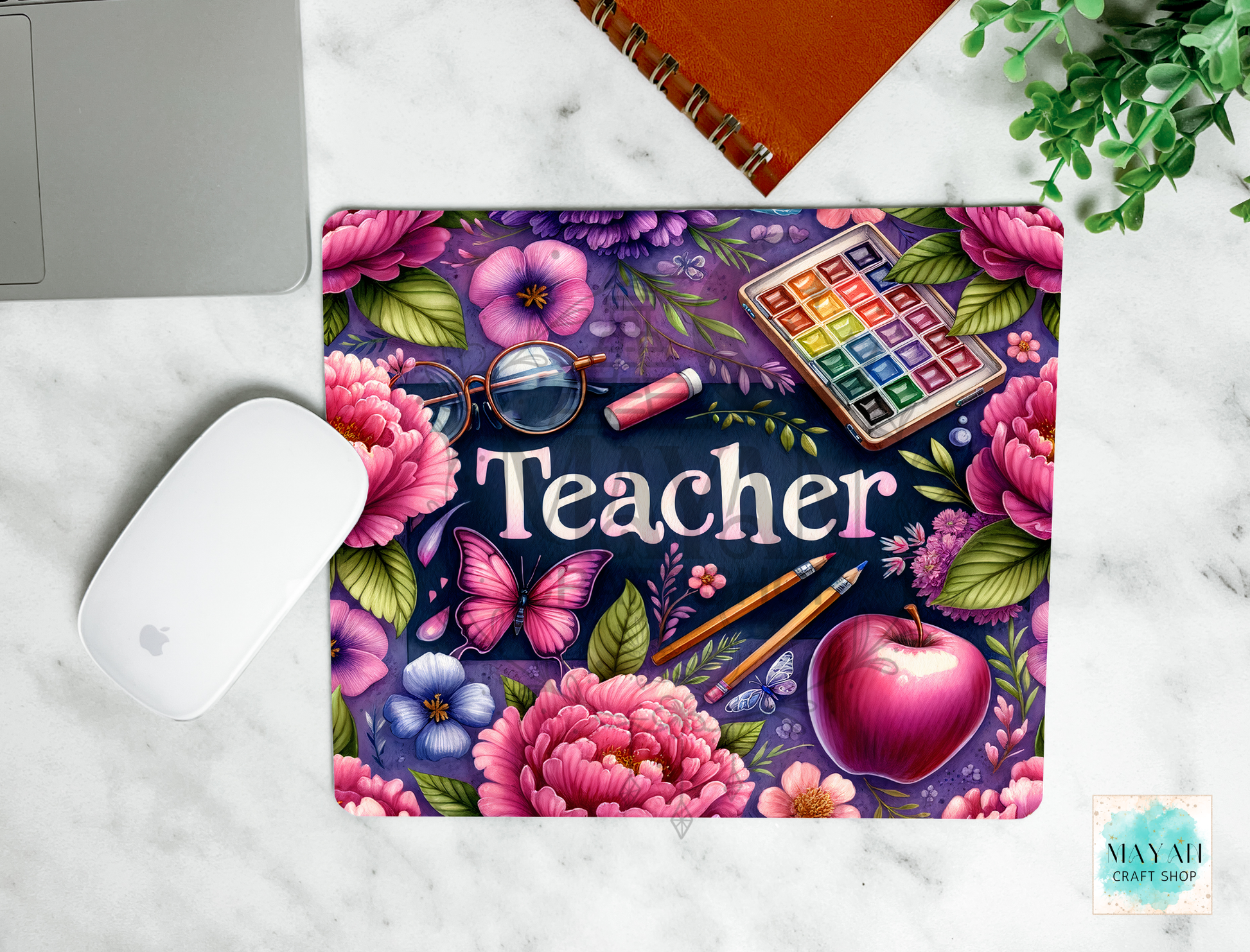 A mouse pad with a colorful floral design and the word 'Teacher' in white, shown on a white marble surface with a laptop mouse, notebook, and plant nearby.