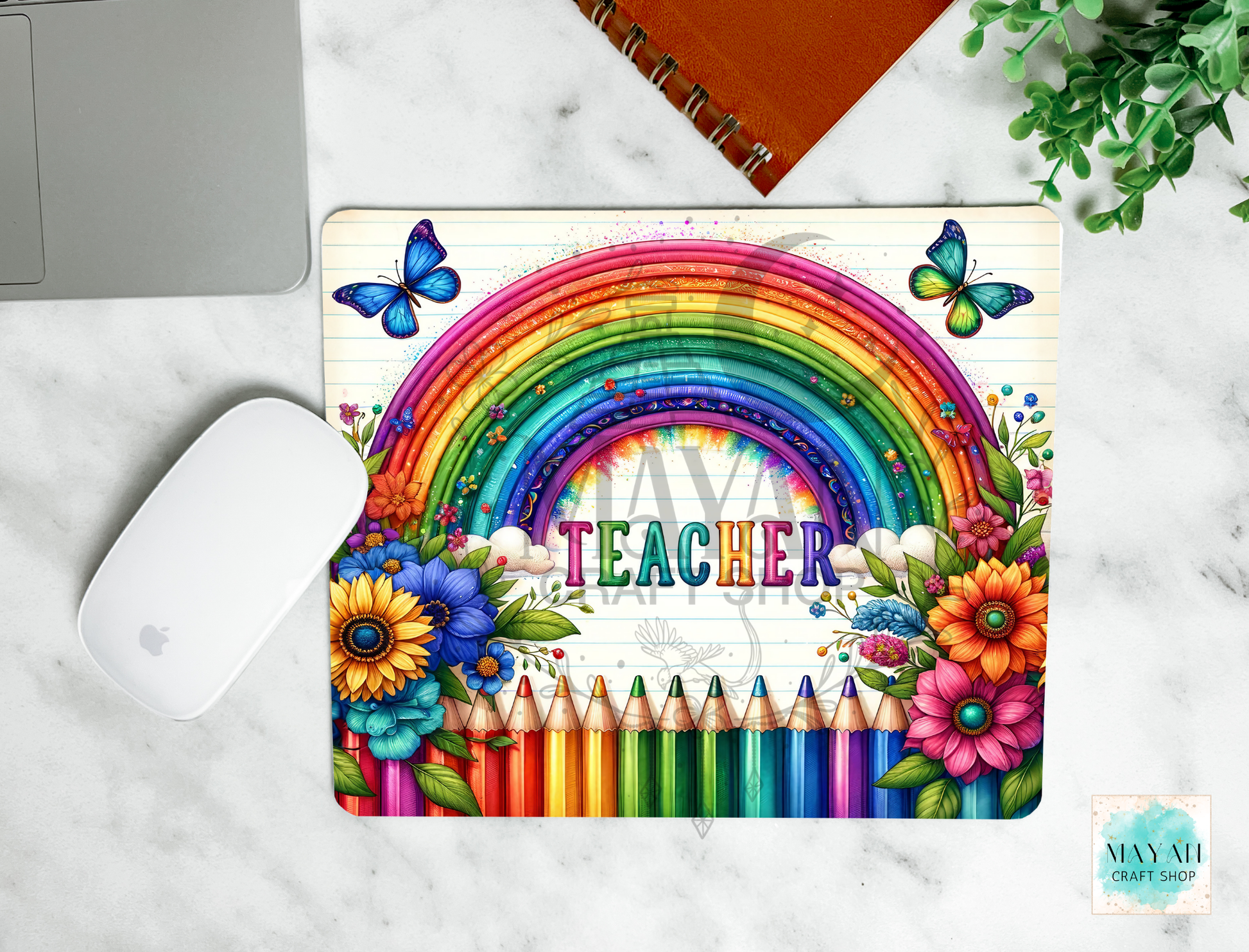 A mouse pad featuring a rainbow, flowers, pencils, butterflies, and the word “TEACHER,” shown in a styled flat-lay on a marble surface with a laptop mouse, notebook, and plant nearby.