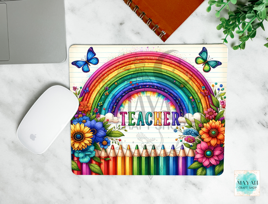 A mouse pad featuring a rainbow, flowers, pencils, butterflies, and the word “TEACHER,” shown in a styled flat-lay on a marble surface with a laptop mouse, notebook, and plant nearby.
