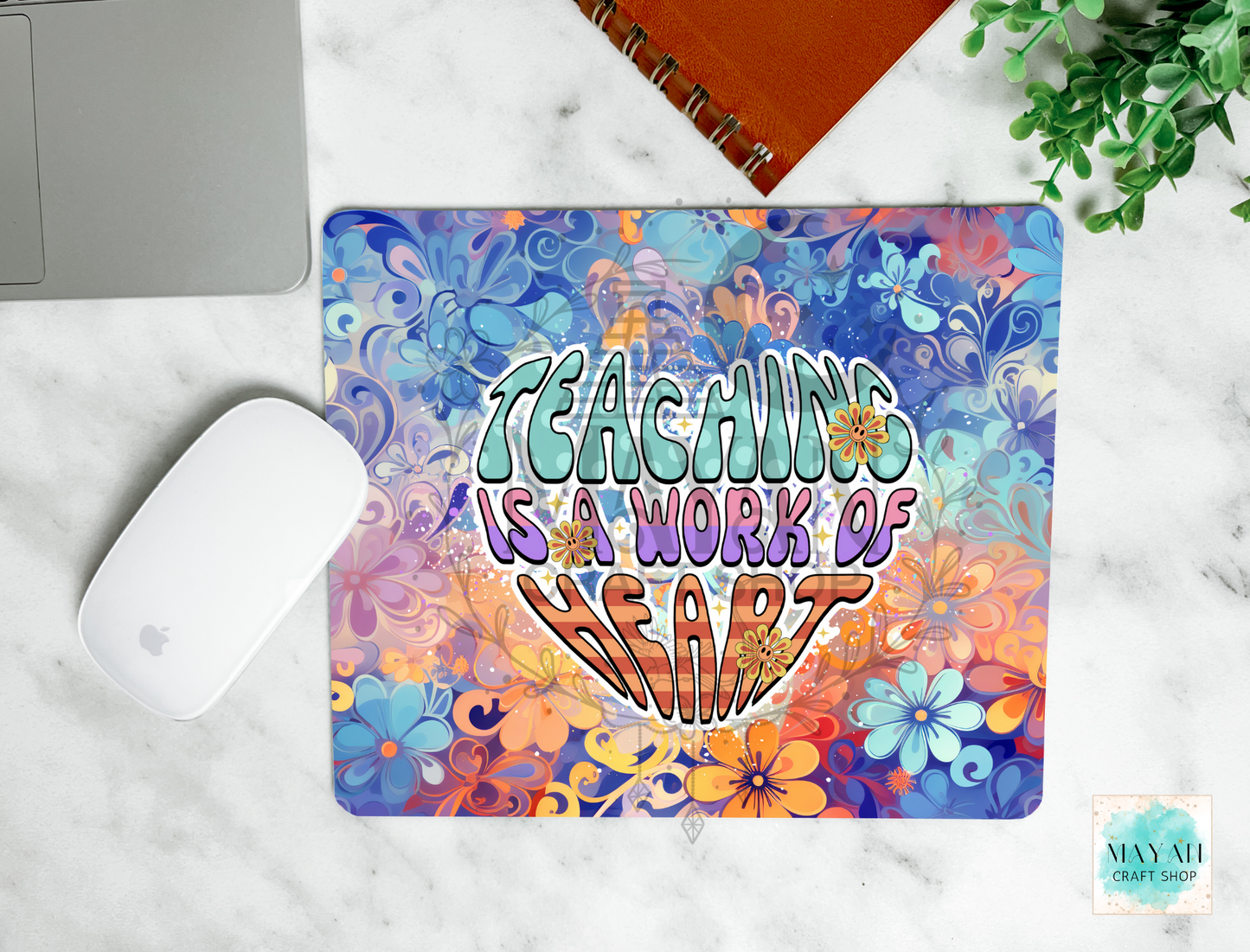 A mouse pad with a colorful retro floral pattern and the text 'TEACHING IS A WORK OF HEART' in stylized lettering, shown on a white marble surface with a laptop, mouse, notebook, and plant nearby.