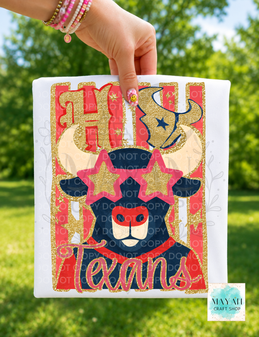 A hand holds a white shirt featuring a Texas-themed bull graphic with gold glitter text, red and navy elements, and a bull head illustration, against a blurred green outdoor background.