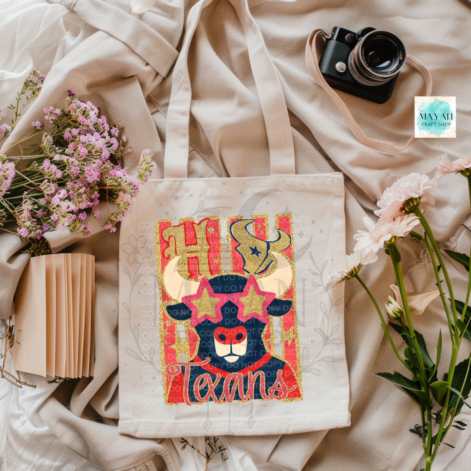 A canvas tote bag with a colorful bull graphic and a text is styled in a flat lay on beige fabric with flowers, a camera, and a notebook. 