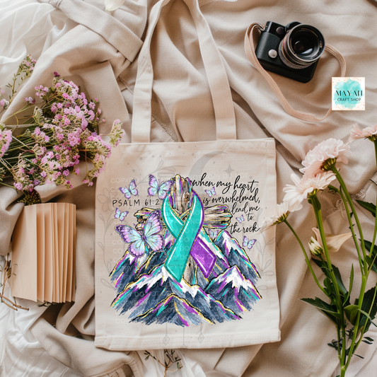 A canvas tote bag with a colorful graphic print featuring a teal and purple awareness ribbon, butterflies, mountains, and script text, styled on beige fabric with flowers, a book, and a camera.