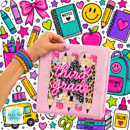 A hand holds a pink shirt featuring three illustrated pencils with leopard print, black glitter, and pink glitter tips, with the words 'first grade' in pink glitter script. The background is a busy school-themed graphic with crayons, notebooks, an apple, smiley face, backpack, and other stationery items.