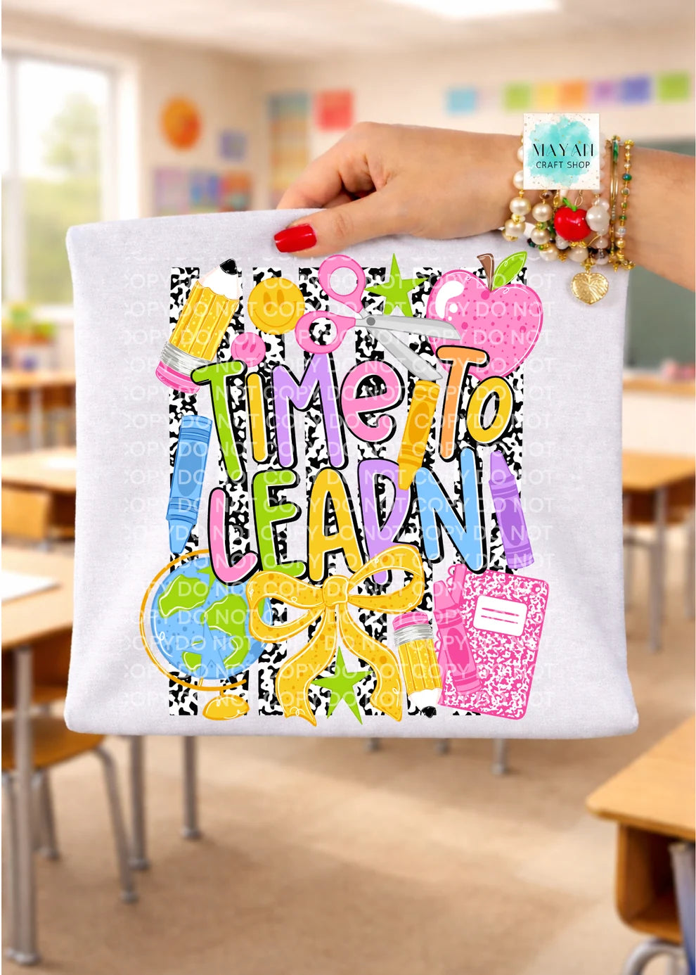 A hand holds a white shirt with a colorful 'Time to Learn' school-themed graphic print, set against a blurred classroom background.