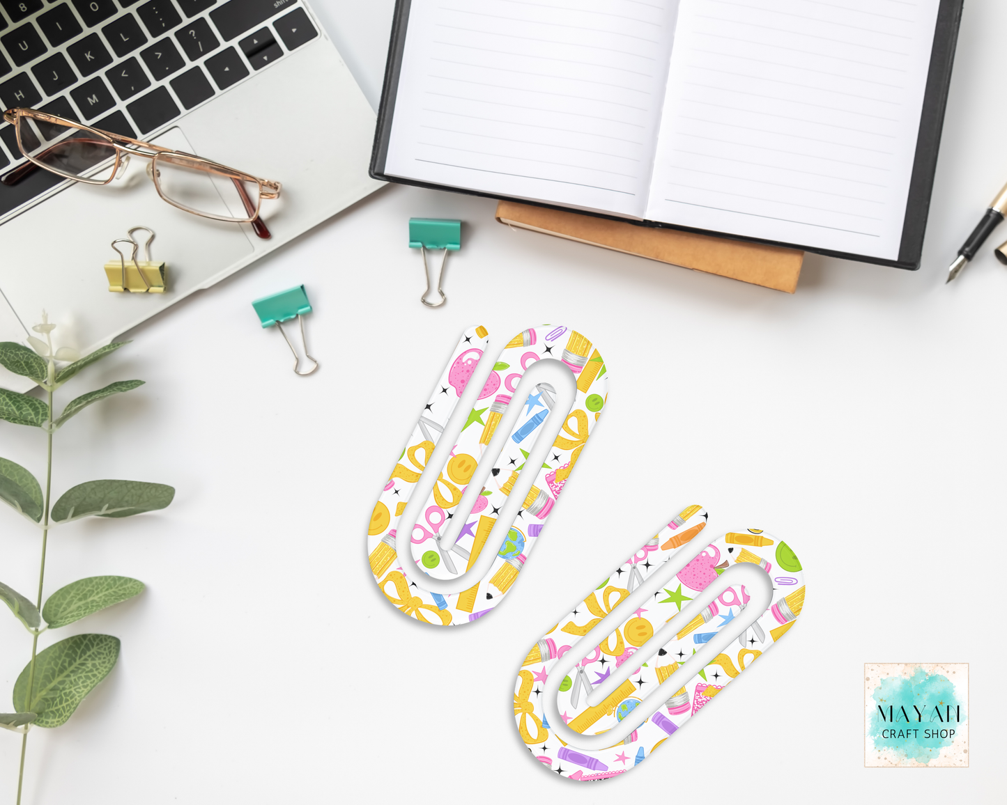 Two paperclip-shaped desk accessories with a colorful pastel pattern of scissors, apples, and stars, displayed on a white desk with office props like a laptop, glasses, binder clips, and an open notebook.