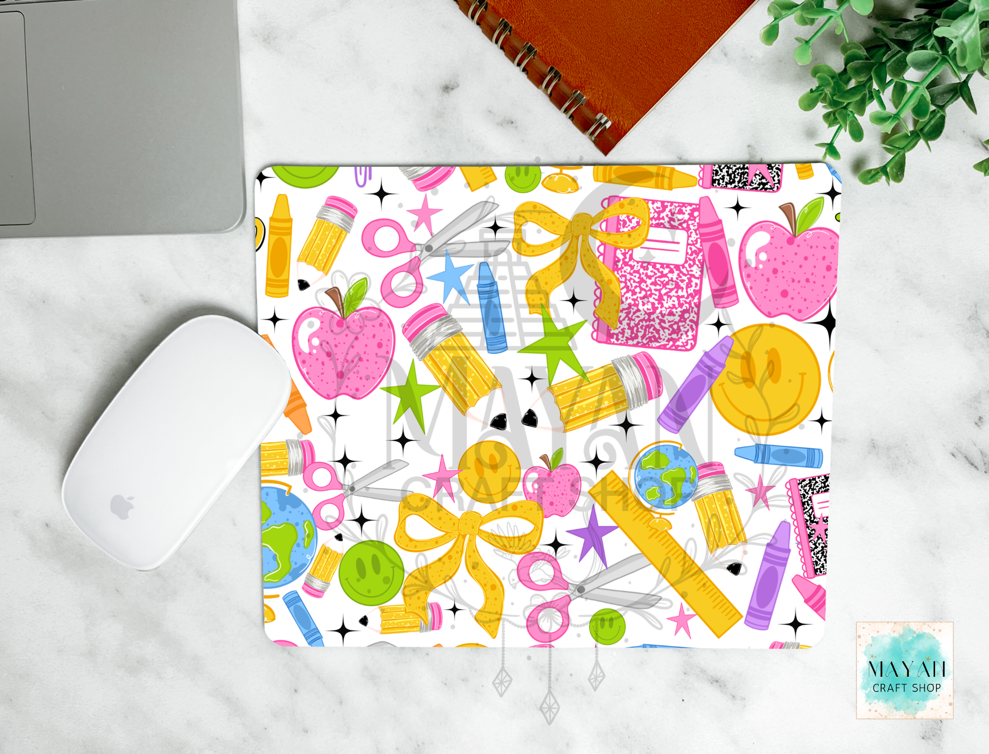 A rectangular mouse pad with a colorful school supply pattern, shown in a styled flat-lay on a white marble surface next to a white computer mouse, a silver laptop corner, an orange spiral notebook, and a green plant. The pattern features pink apples, yellow bows, and various colored pencils, pens, and stars on a white background.