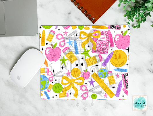 A rectangular mouse pad with a colorful school supply pattern, shown in a styled flat-lay on a white marble surface next to a white computer mouse, a silver laptop corner, an orange spiral notebook, and a green plant. The pattern features pink apples, yellow bows, and various colored pencils, pens, and stars on a white background.