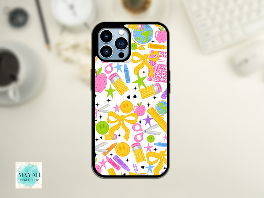 A phone case featuring a colorful school supplies pattern, shown on a phone mockup against a blurred desk background. The pattern includes yellow bows, pink apples, green stars, and blue pencils on a white background.