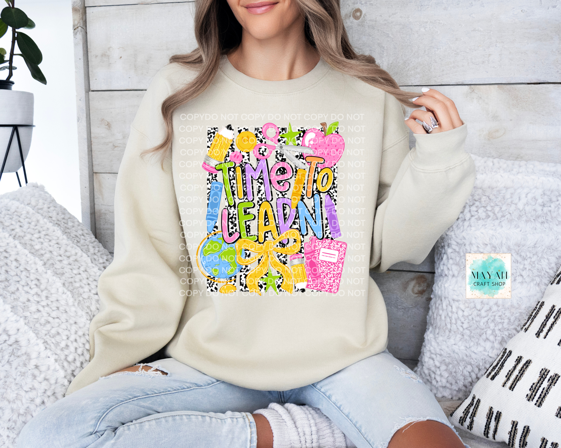 A woman wearing a beige crewneck sweatshirt with a colorful graphic that reads 'TIME TO LEARN' in playful lettering, paired with light blue jeans, sitting on a couch with pillows.