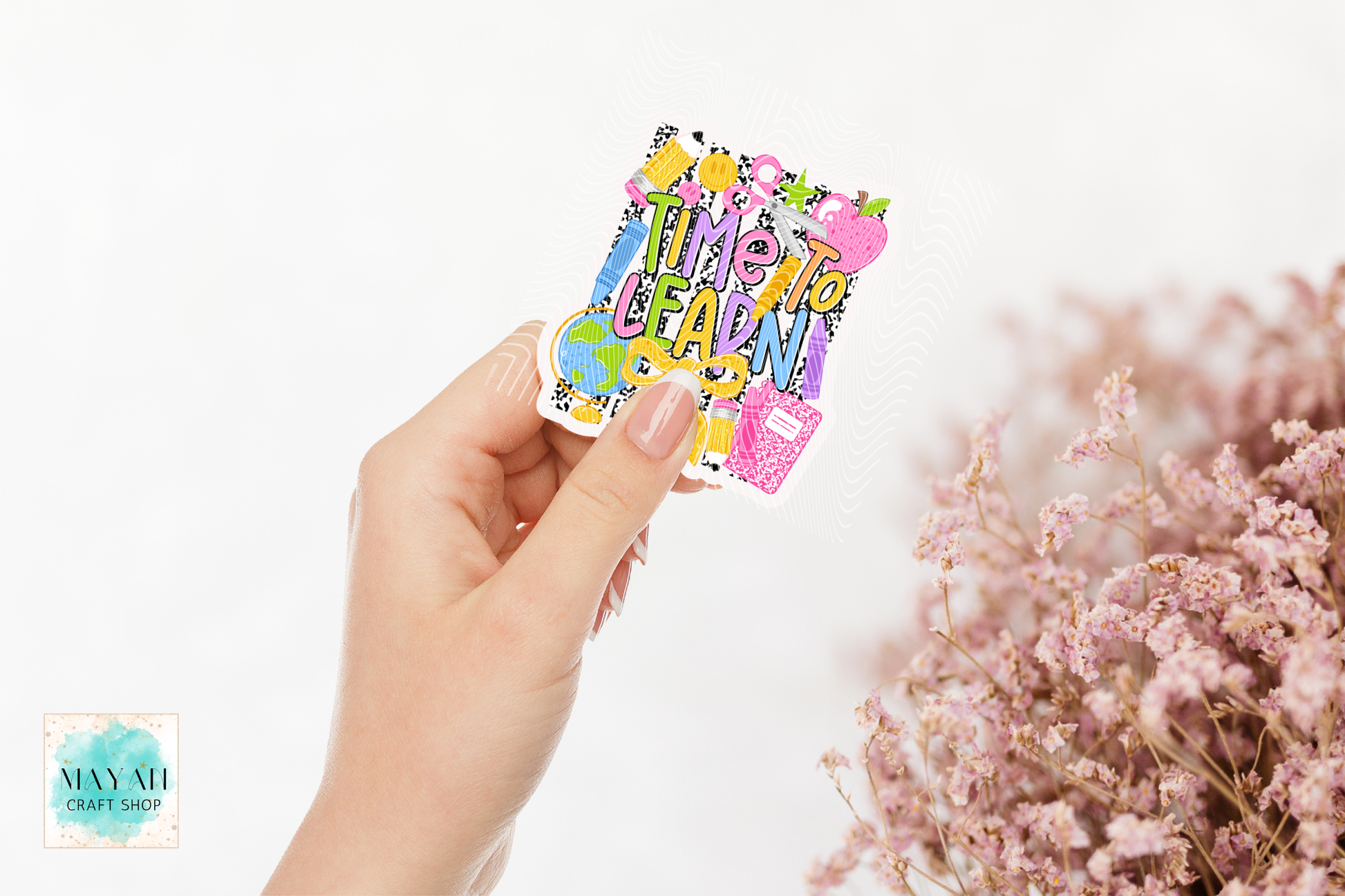 A hand holds a colorful die-cut sticker school patterns, set against a white background with blurred pink flowers on the right.