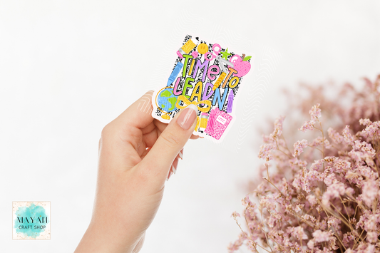 A hand holds a colorful die-cut sticker school patterns, set against a white background with blurred pink flowers on the right.