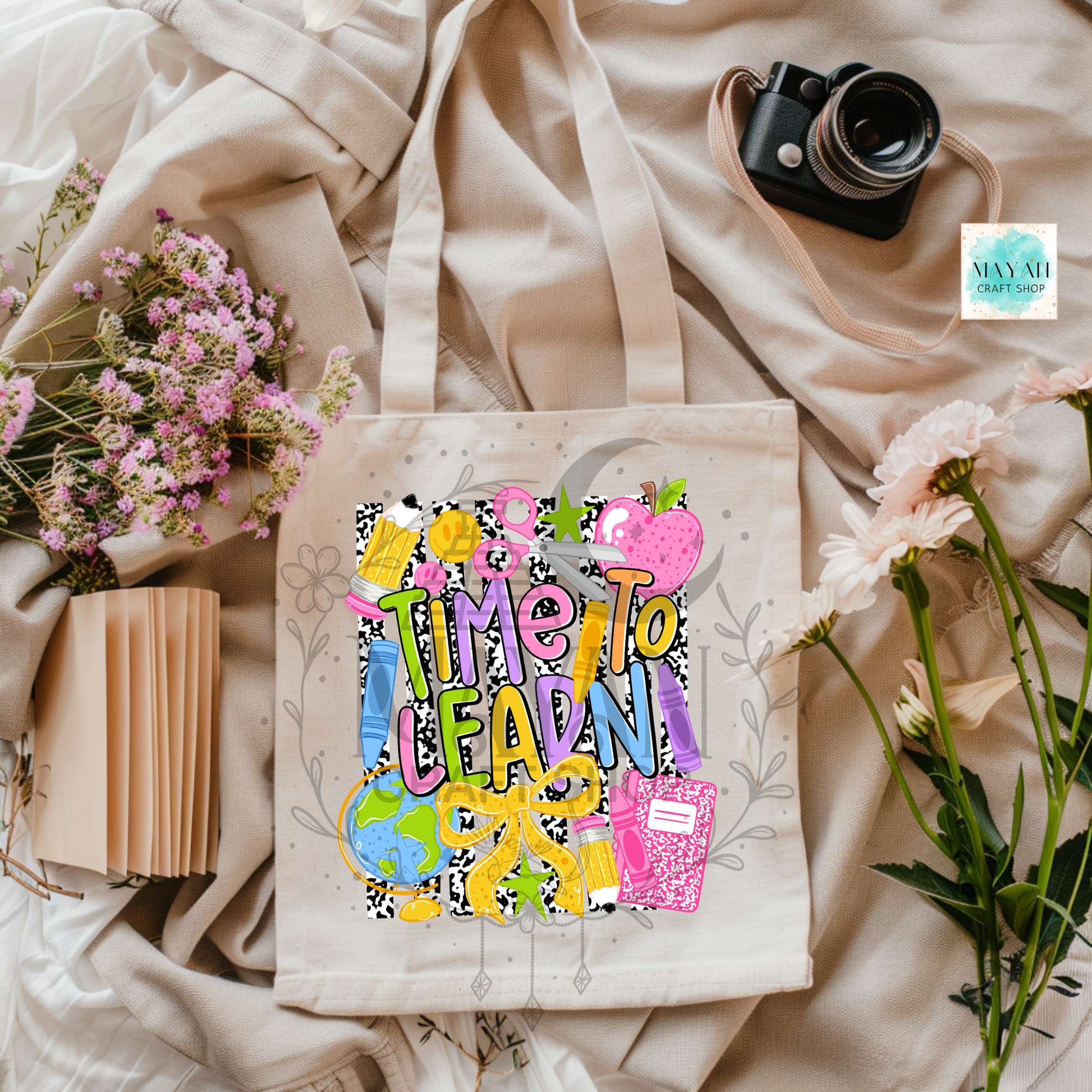 A cream canvas tote bag with a colorful 'TIME TO LEARN!' graphic, styled in a flat lay with beige fabric, pink flowers, a book, and a camera.